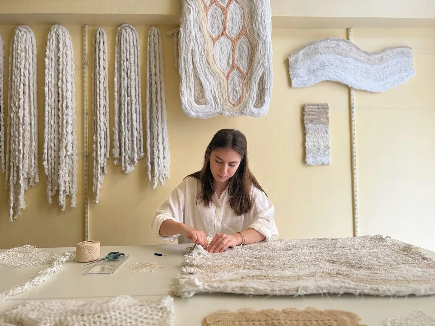 Hara seated at her studio table stitching a textile, with sculptural textiles displayed behind her